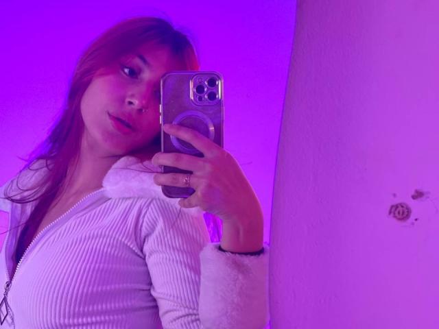 SophieBeaumont with red hair, holding a phone, wearing a white zip-up hoodie, purple background, reflecting in a mirror, showing her tits slightly, smiling, pinkish-purple lighting.