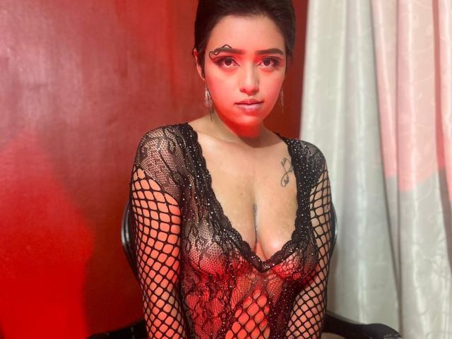 GabbyHot69 with red face makeup, black fishnet top showing her tits, tattoo on right chest, sitting, dark hair, red and white background, looking at camera.