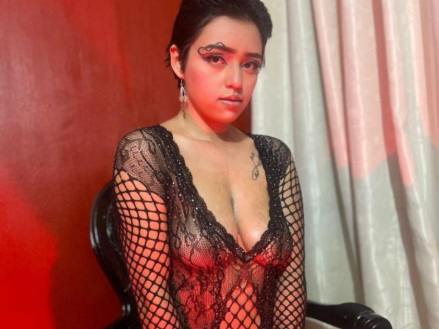GabbyHot69 in a black fishnet dress with a deep cleavage, showing her tits. She has tattoos, dark hair, and red makeup, with a confident expression. Red and white lighting.