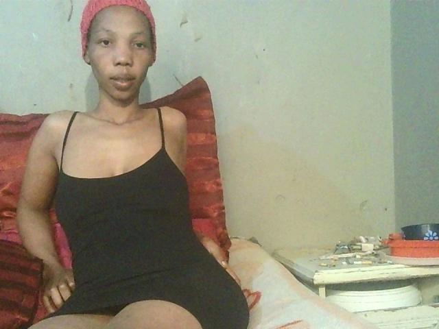 Sexytinny-hot with short red cap, black spaghetti dress, small tits, and dark skin, sitting on bed with red pillows, legs slightly apart, hands on thighs, looking at camera.
