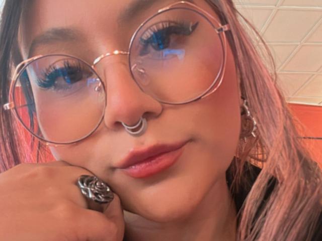 LorraineV with round glasses, nose ring, and hoop earrings, pink lipstick, and a ring, resting her chin on her hand, showing off long eyelashes.