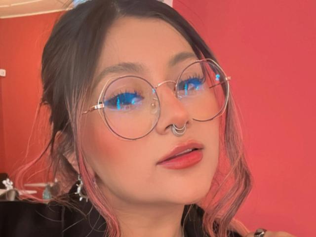 LorraineV with pink-dyed hair, round glasses, blue eyeshadow, red lips, nose ring, and black top, looking slightly to the side.