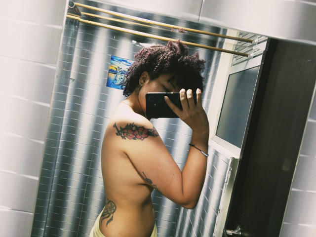 Topless IrySunshine with curly dark hair, holding a phone, showing tattoos on right shoulder and side. Gray tiled bathroom, mirror reflection, greenish-yellow towel. Soft, natural light.
