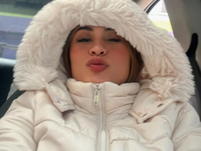 ArianaBellee with pouty lips, wearing a fluffy white hood and a zip-up winter coat, sitting in a car, her face centered in the shot.