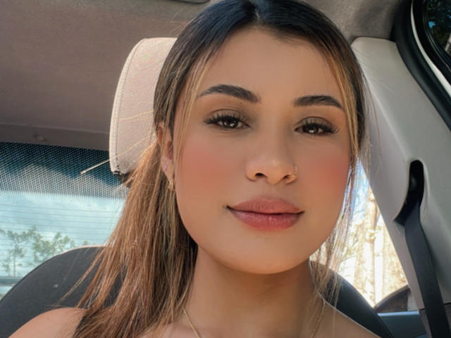 ArianaBellee's face, with brown hair in a ponytail, light skin, thick eyebrows, long lashes, pink lips, and gold earrings, sitting in a car.