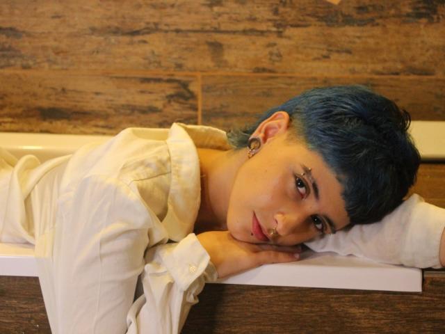 EthanLennox with blue hair, laying on white pillow, wearing white shirt, head resting on hand, wooden background, brown wood wall, ear gauges, serious expression, nose ring.