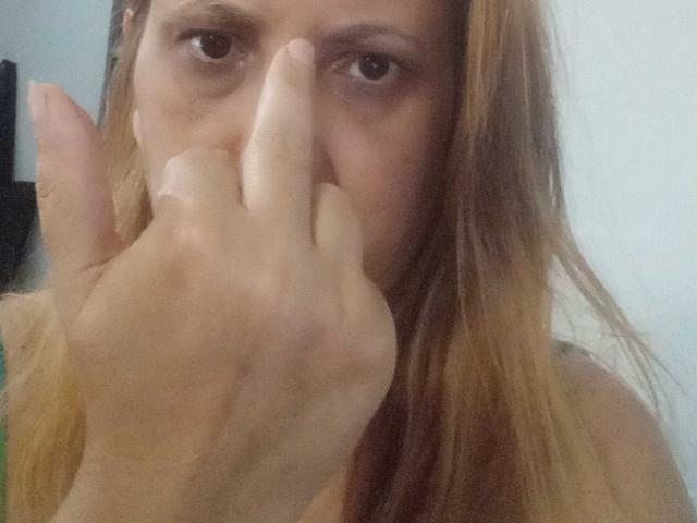 SheronShiedss a les cheveux blond cendré, les yeux marron, et fait un geste de "chut" avec son index sur ses lèvres légèrement entrouvertes, affichant un regard subtil et séduisant..