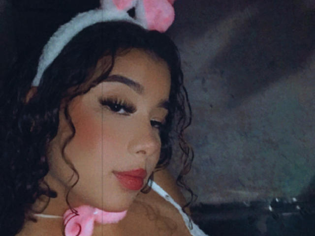 BlossomMia with curly black hair, wearing a white headband with pink bunny ears, and a pink choker, has red lipstick and long eyelashes, looking down with a slight smile.
