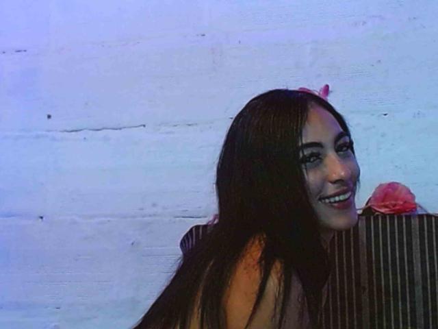 Kendall1storm has long brown hair, dark eyeliner, and is smiling with red lipstick, leaning on a striped object, wearing a pink flower in her hair.