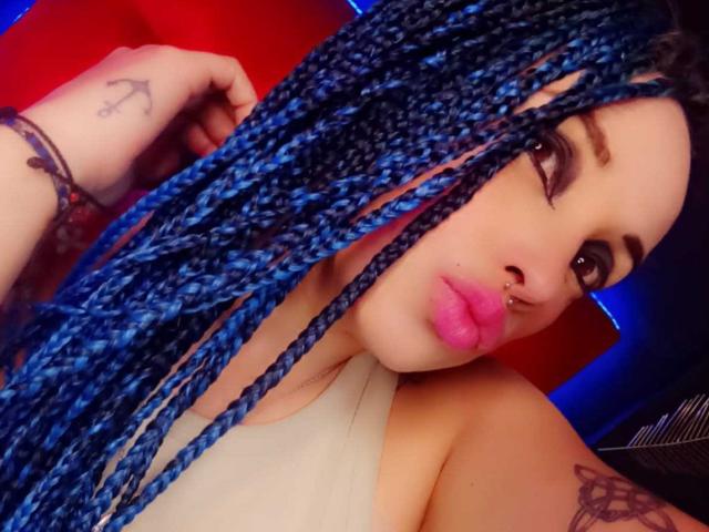 Dalila Musa, com tranças azuis, lábios rosa, piercing no nariz, tatuagem na mão e tatuagem no braço, vestindo uma camiseta branca, olhando para a câmera com uma expressão emburrada..