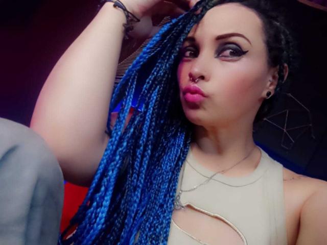 Dalila Musa, met blauwe vlechten, roze lippenstift en zwarte eyeliner, zet haar volle lippen op een zuur gezicht. Ze draagt een beige top met uitsparingen, waardoor haar borsten zichtbaar zijn. Haar arm is omhoog..