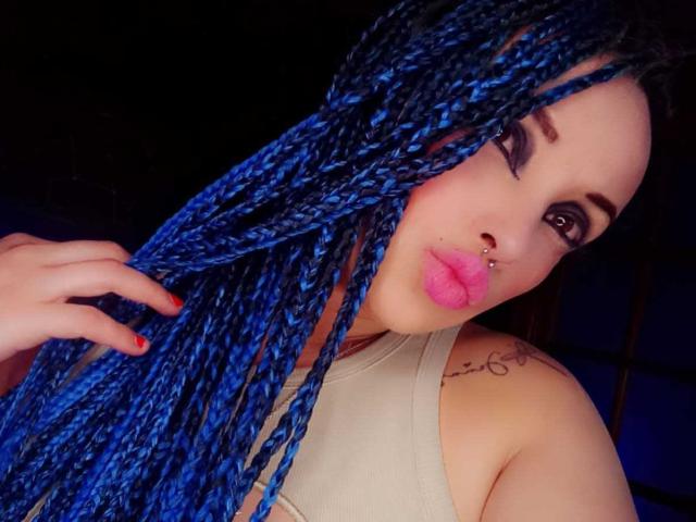 Dalila Musa, com tranças azuis vibrantes, batom rosa e um piercing no nariz, segurando o cabelo, mostrando uma tatuagem no ombro, vestindo uma blusa bege, olhando para a câmera..
