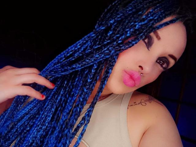 DalilaMusa with blue braids, pink lipstick, nose ring, and tattoo on shoulder, holding her hair, wearing a beige top, with red nails, and dark eye makeup.
