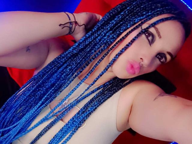 Dalila Musa, com tranças azuis, lábios rosados e cheios, um piercing no nariz e tatuagens, vestindo uma blusa branca, tirando uma selfie, mostrando seus seios e braços. Fundo azul e vermelho..