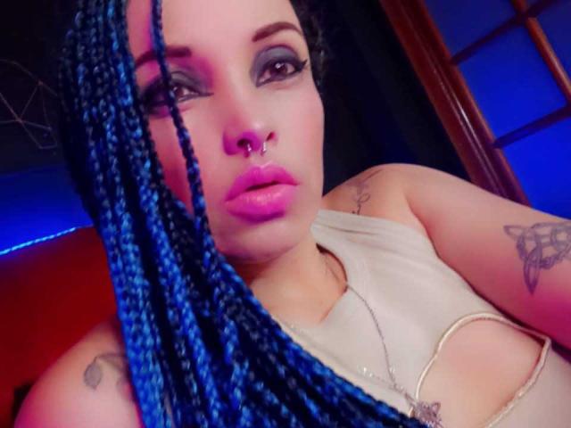 Dalila Musa, met blauwe vlechten, roze lippen, een neusring en tatoeages, draagt een witte top en kijkt naar de camera met donkere, smoky oogmake-up en een lichte frons..