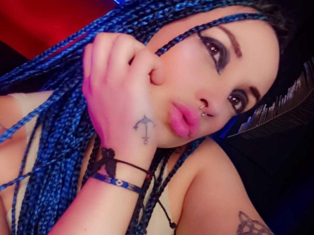 Dalila Musa, con le trecce blu, le labbra rosa, il piercing al naso e i tatuaggi, si tiene la guancia, indossa un braccialetto nero, ha un aspetto sexy e un bel sedere..