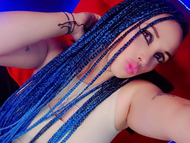 Dalila Musa, com tranças azuis, lábios rosados e cheios, piercing no nariz e tatuagens, vestindo uma blusa branca que mostra seus seios e tatuagens nos braços, com um fundo vermelho..