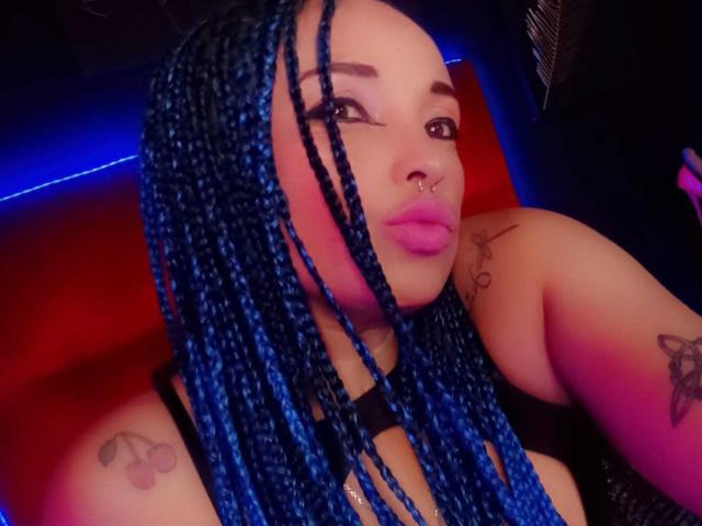 Dalila Musa, con trecce blu, labbra rosa, un anello al naso, tatuaggi sulla spalla e sul petto, indossa una maglietta nera, in una stanza poco illuminata con luci blu e rosse..
