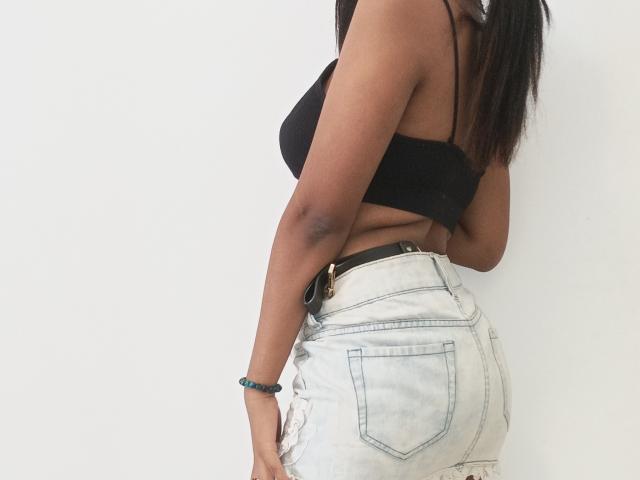 MaliciaQueen, de piel oscura, con un top negro ajustado que deja ver sus pequeños pechos, y pantalones cortos vaqueros blancos desgastados, lleva un cinturón negro y una pulsera azul, y está de pie de perfil..