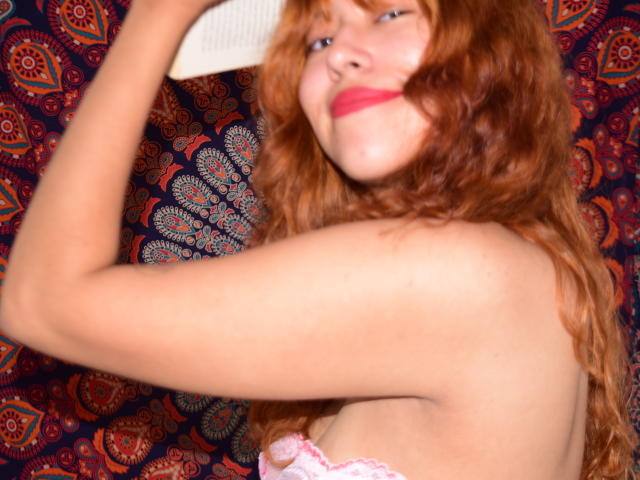 GigimonRamia met rood krullend haar, een roze kant beha, en rode lippenstift, die lichtjes glimlacht, met een arm omhoog, een wit object vasthoudend, waarbij haar blote schouder en bovenkant van haar borst zichtbaar zijn..