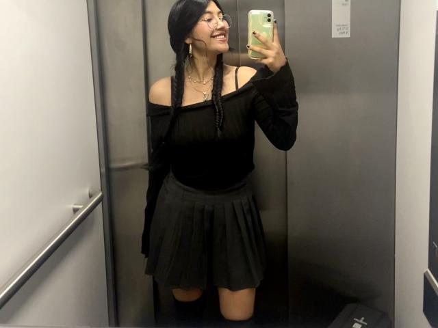 Elizaa with dark braids, off-shoulder black top, and black skirt, taking a selfie in a elevator, smiling, showing her tits and ass, holding a green phone.