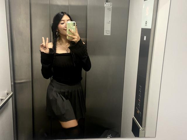 Elizaa in un ascensore, indossa una maglietta nera con una spalla scoperta, una gonna nera, ha i capelli lunghi e neri, fa il segno della pace, tiene un telefono in mano, ha un piccolo tatuaggio sul collo, ha un seno di media grandezza e indossa calze nere alte fino alla coscia..