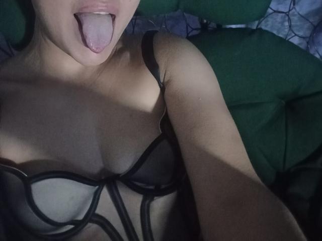 EvaAlferash's tongue is out, showing her pink tongue. She's wearing a black, sheer, strapless top with visible boobs and black straps. Her skin is light.