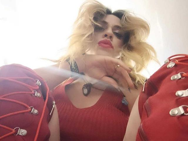 PaulettWatsoon with blonde hair, red lipstick, red lace-up sleeves, and a red ribbed top, looking down at the camera with a hand near their chin.