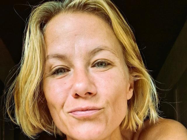 xcYola-hot has blonde, shoulder-length hair, light skin, green eyes, and a subtle smile, showing slight wrinkles and a natural, relaxed look. Her small nose and light pink lips are visible.