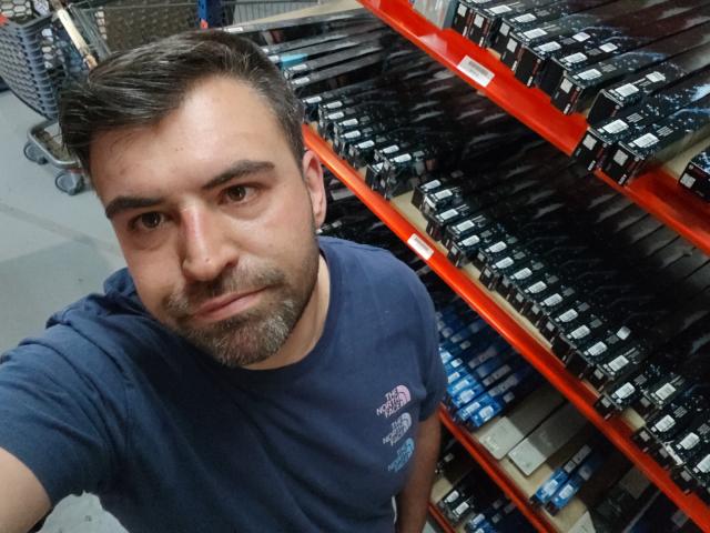 Vio69x, a bearded man with dark hair, wearing a navy blue shirt, standing in a store with red shelves filled with black boxes, looking at the camera.