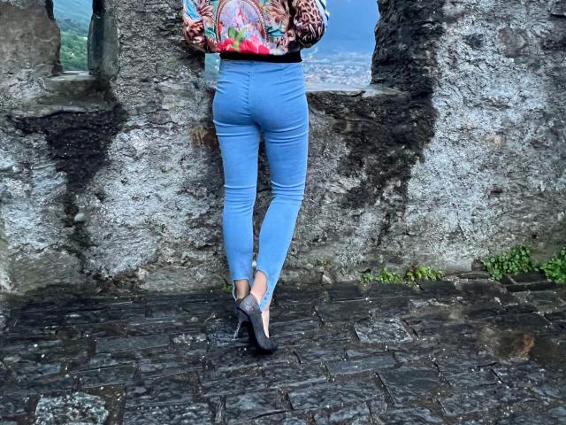 LaPresuntuosa-hot è in piedi di fronte alla telecamera con il sedere rivolto, indossa jeans blu, tacchi alti neri e uno zaino colorato, con alle spalle un muro di pietra grezza. Il suo sedere è tonico e attraente..