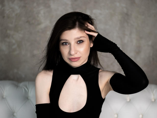 NanceyLove with pale skin, dark brown hair, and blue eyes, wearing a black cutout dress and arm sleeve, touching her head, sitting on a white tufted sofa.