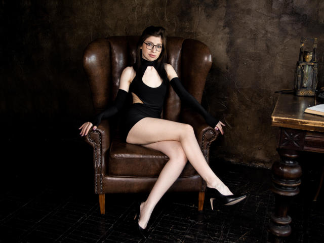 NanceyLove, with glasses, sitting on a brown leather chair, wearing a black cutout dress, black arm sleeves, and black heels, showing her pale legs and small tits.