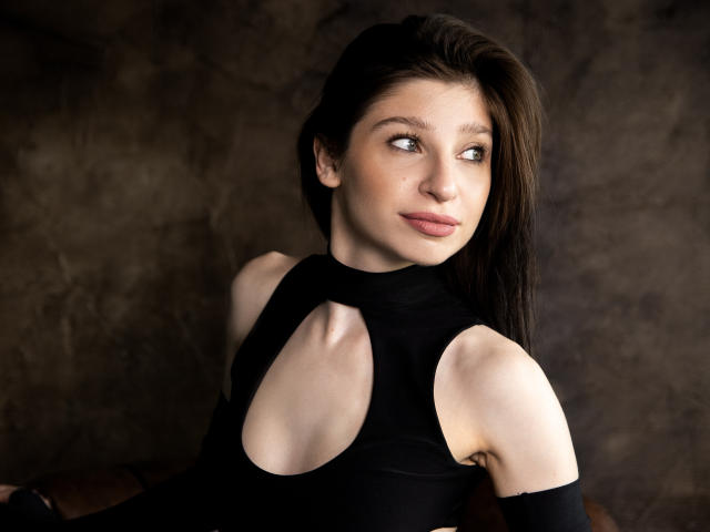 NanceyLove with pale skin, dark brown hair, and subtle makeup, wearing a black top with cutout shoulders, looking to the side with a slight smile.