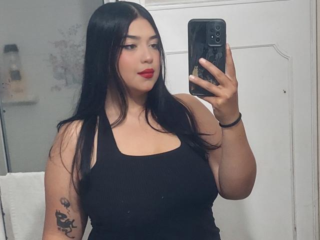 Jennadavon with long black hair, red lipstick, black tank top, holding phone, showing tattoo on left arm, taking mirror selfie, wearing black bracelet, standing in bathroom.