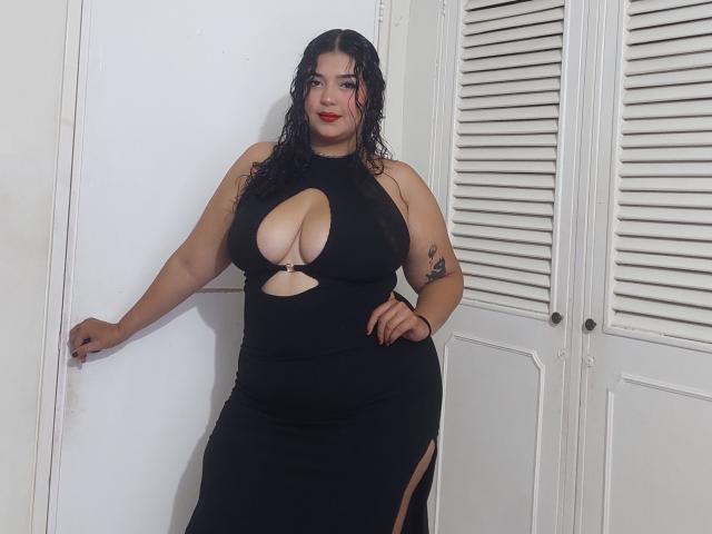 Jennadavon in a black dress with a large cutout showing her tits, standing with wet black hair, red lipstick, tattoo on her right arm, and a confident smile.