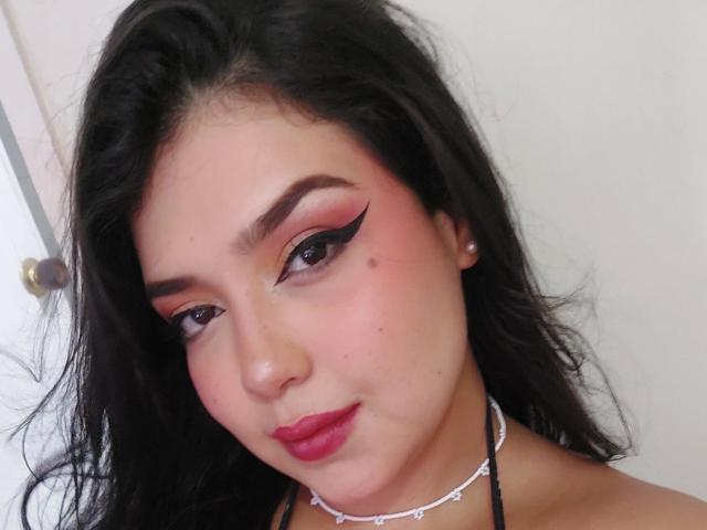 Jennadavon with black hair, pink blush, sharp black eyeliner, red lipstick, small pearl earrings, and a white choker necklace, looking at the camera with a slight smile.