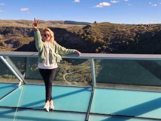 FreankyQueen staat met een vrede-teken, een groene jas, een wit shirt, zwarte broek en witte sneakers op een glazen balkon, met blond haar, een zonnebril en een schattige achterkant..
