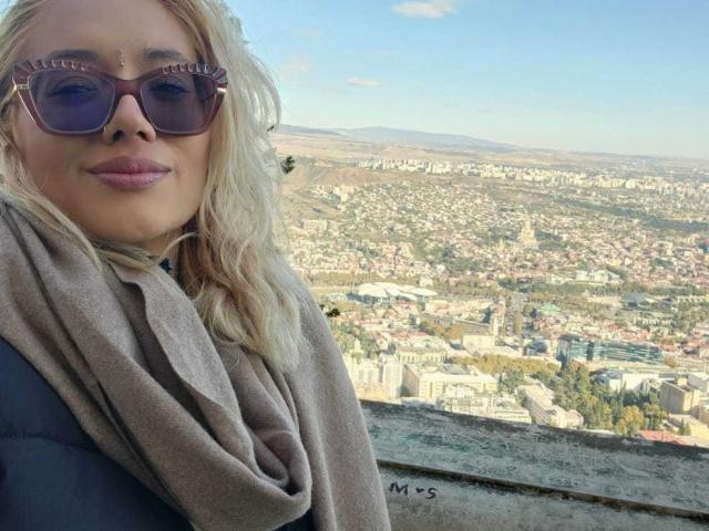 FreankyQueen with blonde hair, wearing sunglasses and a grey scarf, has puffy pink lips and is looking down at a cityscape from a high viewpoint.