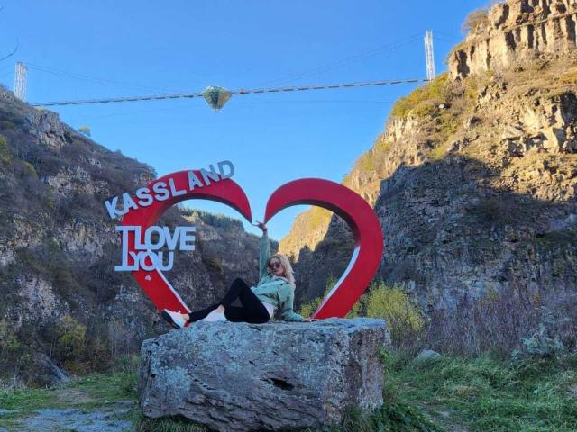 FreankyQueen is sitting on a rock with a red heart and "Kassland Love" sign, wearing a green shirt and black pants, in a rocky mountain valley.