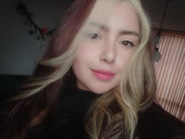 Aradiia, con capelli biondi e rosso scuro, labbra rosa e una maglia nera, sorride dolcemente in una foto, mostrando il suo viso e la parte superiore del petto..