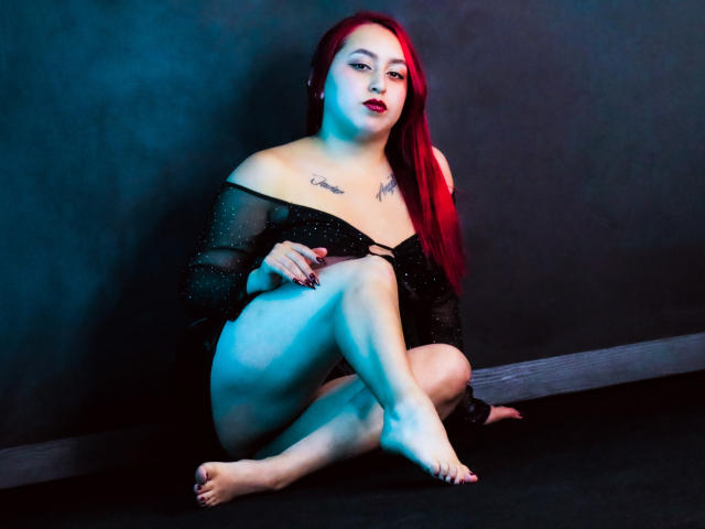 ElenaWesteros met rood haar, een zwarte doorschijnende top en zwarte shorts, zittend op de vloer, benen gekruist, tatoeages op de borst, blauwe en rode verlichting, donkere achtergrond, rode lipstick, nagels gelakt..