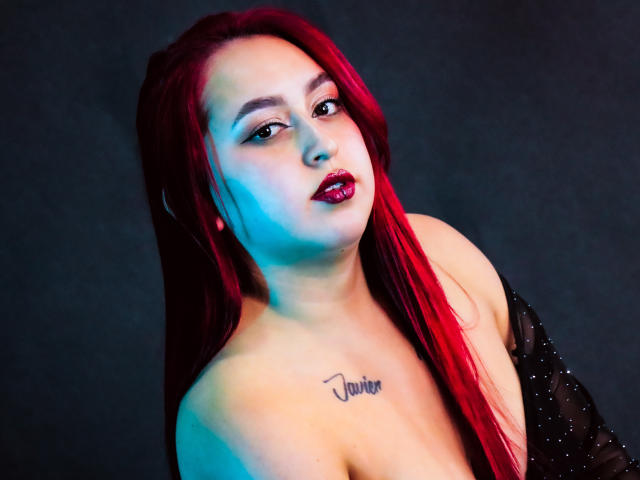 ElenaWesteros con capelli rossi, spalle scoperte, top nero trasparente, illuminazione blu e rossa, tatuaggio sul petto, rossetto rosso, che guarda verso l'alto con le labbra leggermente dischiuse..
