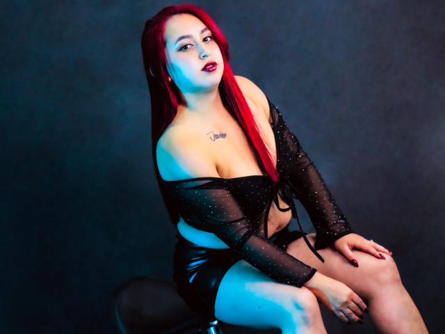 ElenaWesteros, con cabello rojo, una blusa negra transparente y pantalones cortos negros, sentada en un taburete, mostrando sus pechos, con un tatuaje en el pecho, iluminación azul y roja, y una expresión de confianza..