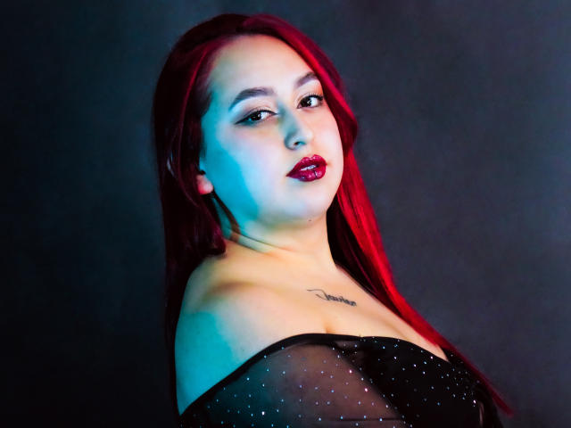 ElenaWesteros, com cabelo ruivo, batom vermelho, blusa preta transparente, pequena tatuagem no peito, olhando para a câmera com iluminação azul e vermelha. Seios parcialmente visíveis através da blusa transparente..