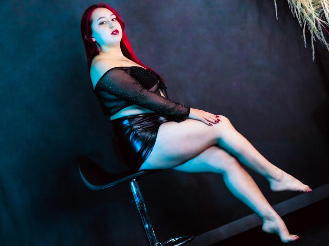 ElenaWesteros con i capelli rossi, che indossa una maglia nera trasparente e pantaloncini neri lucidi, seduta su uno sgabello, con le gambe incrociate, piedi nudi, sfondo scuro, illuminazione blu e rossa, sexy, sicura di sé..