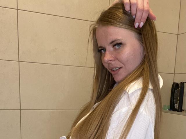 JollyKissa with long blonde hair, white shirt, holding her hair up with one hand, smiling at the camera, blue eyes, in a bathroom. Hand on her head, looking sexy.