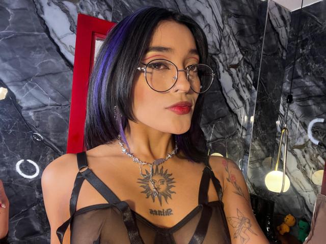SalomeMille with black hair, glasses, red lipstick, and a sun tattoo on her chest, wearing a sheer black top, showing small tits and tattoos, in a room with dark marble.