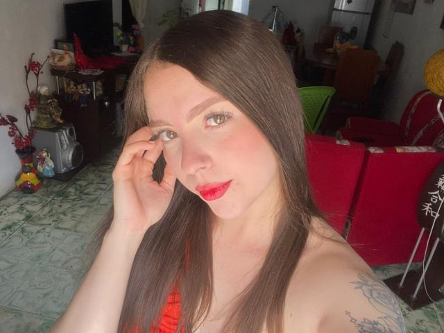 LouiseBallen with long brown hair, green eyes, red lipstick, and a tattoo on her right arm, touching her face with a slight smile, showing her bare shoulders.