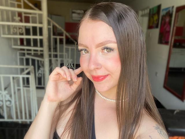 LouiseBallen with long brown hair, red lipstick, and a pearl necklace, touching her nose with a slight smile, showing tattoos on her right shoulder.